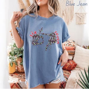May include: Blue jean colored t-shirt with a skeleton hand graphic. The graphic shows two skeleton hands forming the number six and seven with Christmas lights and the words "Six" and "Seven" in red.