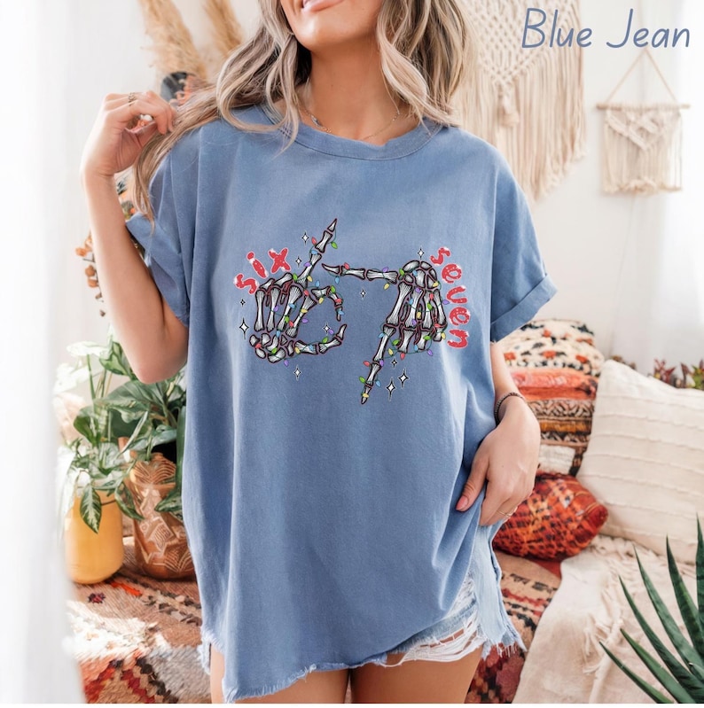May include: Blue jean colored t-shirt with a skeleton hand graphic. The graphic shows two skeleton hands forming the number six and seven with Christmas lights and the words "Six" and "Seven" in red.