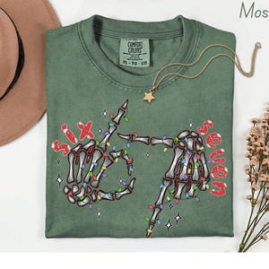 May include: A moss green t-shirt with a skeleton hand graphic, strung with colorful Christmas lights, and the words "Six" and "Seven" in red. A gold star pendant necklace and a brown hat are also visible.