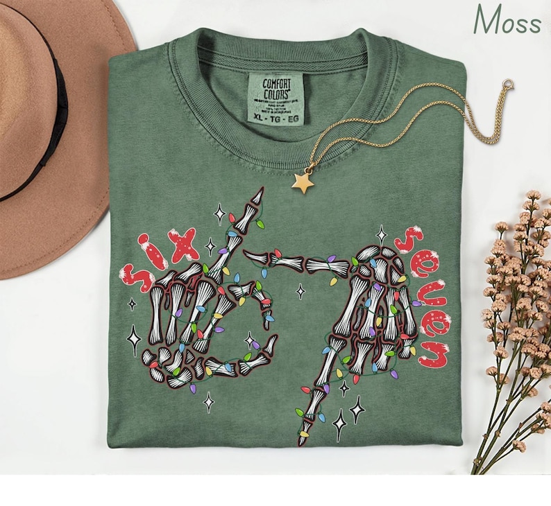 May include: A moss green t-shirt with a skeleton hand graphic, strung with colorful Christmas lights, and the words "Six" and "Seven" in red. A gold star pendant necklace and a brown hat are also visible.