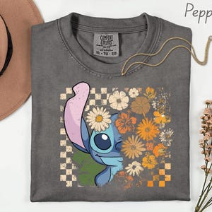 Puede incluir: Una camiseta gris Comfort Colors con un gr&aacute;fico de Stitch y un dise&ntilde;o floral. El dise&ntilde;o incluye un patr&oacute;n de tablero de ajedrez, margaritas y flores naranjas y amarillas. Un collar dorado est&aacute; drapeado sobre la camisa.