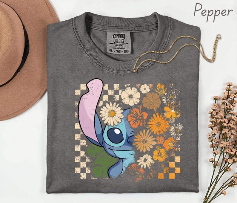 Puede incluir: Una camiseta gris Comfort Colors con un gr&aacute;fico de Stitch y un dise&ntilde;o floral. El dise&ntilde;o incluye un patr&oacute;n de tablero de ajedrez, margaritas y flores naranjas y amarillas. Un collar dorado est&aacute; drapeado sobre la camisa.