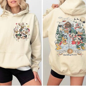 May include: A cream-colored hoodie with a Christmas-themed Mickey Mouse print on the front and a larger print on the back featuring Mickey Mouse and friends at a winter wonderland scene. The back print includes the text "Monster's Inc. In The Works Walt Disney World".