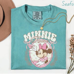 Puede incluir: Camiseta verde mar con un gr&aacute;fico de Minnie Mouse. El dise&ntilde;o incluye el texto "Minnie Mouse", un lazo rosa y una ilustraci&oacute;n de dibujos animados de Minnie Mouse. Un collar con colgante de estrella dorada est&aacute; sobre la camisa.