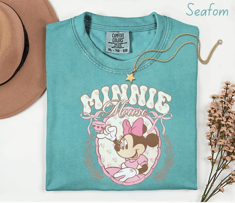 Puede incluir: Camiseta verde mar con un gr&aacute;fico de Minnie Mouse. El dise&ntilde;o incluye el texto "Minnie Mouse", un lazo rosa y una ilustraci&oacute;n de dibujos animados de Minnie Mouse. Un collar con colgante de estrella dorada est&aacute; sobre la camisa.