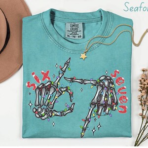 May include: Seafoam green Comfort Colors t-shirt with a skeleton hand graphic. The graphic features two skeleton hands with colorful Christmas lights and the words "Six" and "Seven" in red. A gold necklace with a star pendant is on the shirt.