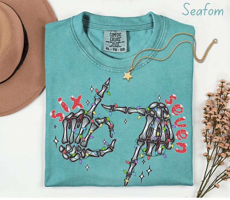 May include: Seafoam green Comfort Colors t-shirt with a skeleton hand graphic. The graphic features two skeleton hands with colorful Christmas lights and the words "Six" and "Seven" in red. A gold necklace with a star pendant is on the shirt.