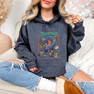 Pu&ograve; includere: Felpa con cappuccio grigio scuro con la scritta "Zootopia" sopra una grafica colorata dei personaggi del film d'animazione. La grafica include una volpe, un coniglio e altri animali. La felpa ha un cappuccio con coulisse e una tasca frontale.