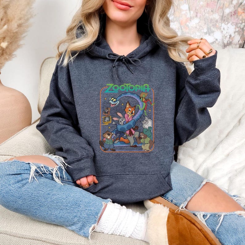 Pu&ograve; includere: Felpa con cappuccio grigio scuro con la scritta "Zootopia" sopra una grafica colorata dei personaggi del film d'animazione. La grafica include una volpe, un coniglio e altri animali. La felpa ha un cappuccio con coulisse e una tasca frontale.