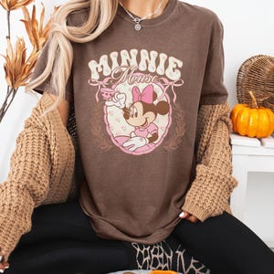 Puede incluir: Camiseta marr&oacute;n con un gr&aacute;fico de Minnie Mouse. El dise&ntilde;o incluye el texto "Minnie Mouse" en una fuente decorativa, junto con una ilustraci&oacute;n de Minnie Mouse con un vestido rosa. Es una prenda informal.