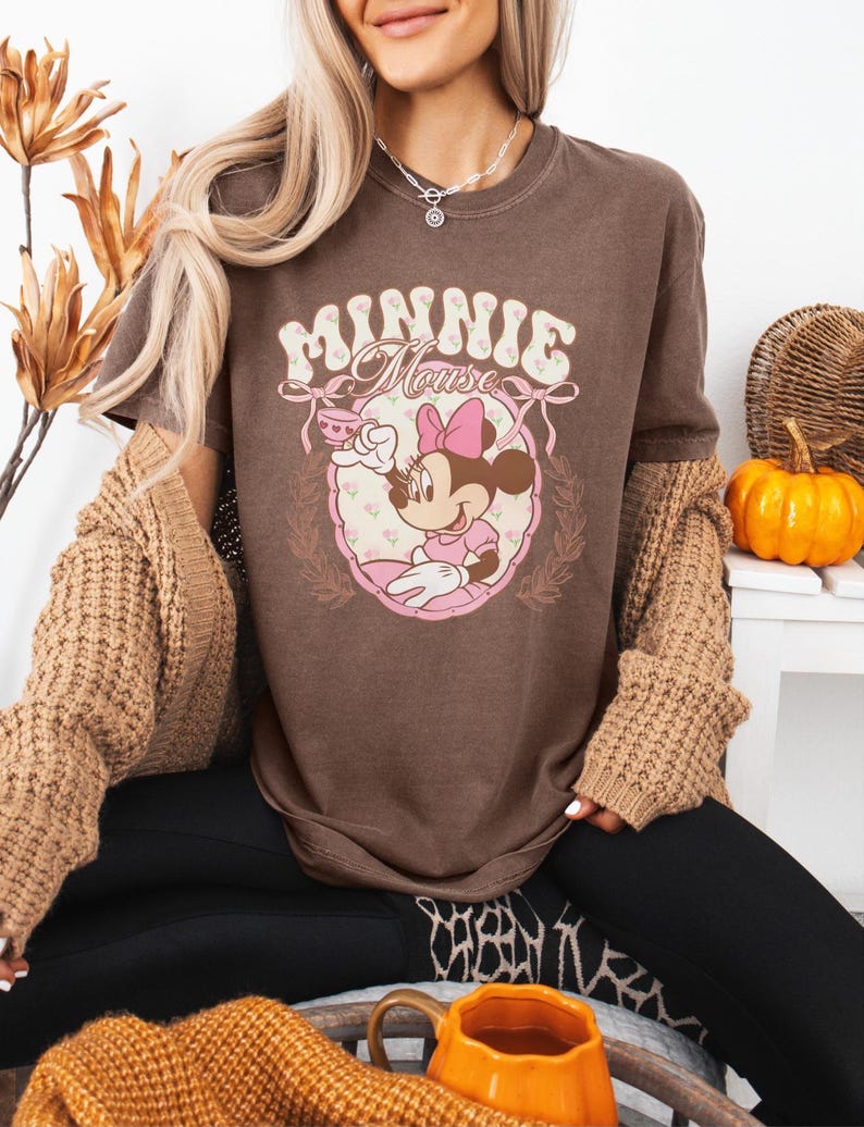 Puede incluir: Camiseta marr&oacute;n con un gr&aacute;fico de Minnie Mouse. El dise&ntilde;o incluye el texto "Minnie Mouse" en una fuente decorativa, junto con una ilustraci&oacute;n de Minnie Mouse con un vestido rosa. Es una prenda informal.