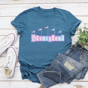 May include: A teal blue t-shirt with a graphic of the word "Disneyland" in colorful blocks with flags above the letters. The shirt is laid out on a white wooden surface with a pair of white canvas shoes and blue jeans.