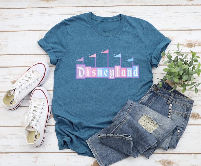May include: A teal blue t-shirt with a graphic of the word "Disneyland" in colorful blocks with flags above the letters. The shirt is laid out on a white wooden surface with a pair of white canvas shoes and blue jeans.