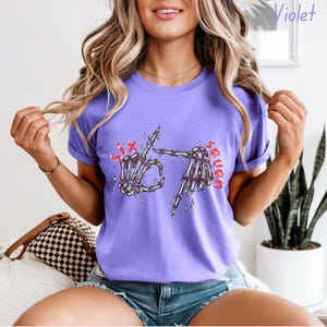 May include: A violet t-shirt with rolled sleeves featuring a skeleton hand graphic. The graphic includes the words "Six" and "Seven" with festive lights and star accents. The shirt is a solid color.