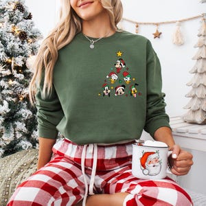 Pode incluir: Uma camisa de l&atilde; verde com um design de &aacute;rvore de Natal com v&aacute;rios personagens da Disney, incluindo Mickey Mouse, Donald Duck, Pateta e Pluto. A camisa de l&atilde; est&aacute; sendo usada por uma pessoa sentada em uma manta xadrez vermelha e branca, segurando uma caneca branca com um design de Papai Noel.