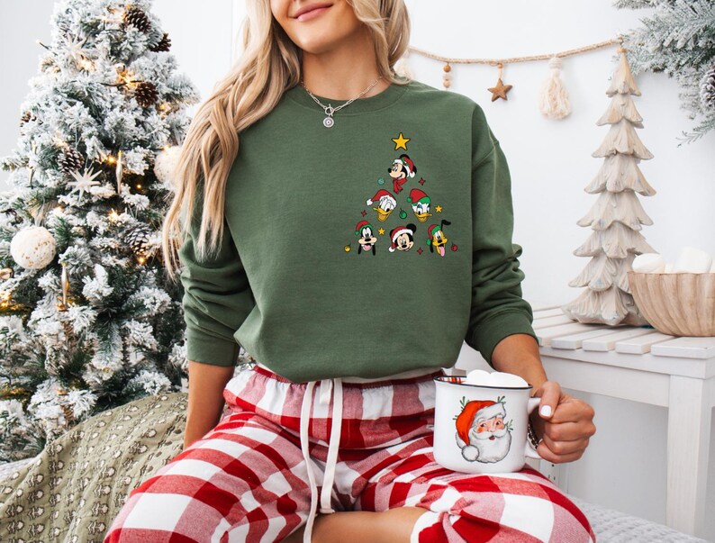 Pode incluir: Uma camisa de l&atilde; verde com um design de &aacute;rvore de Natal com v&aacute;rios personagens da Disney, incluindo Mickey Mouse, Donald Duck, Pateta e Pluto. A camisa de l&atilde; est&aacute; sendo usada por uma pessoa sentada em uma manta xadrez vermelha e branca, segurando uma caneca branca com um design de Papai Noel.