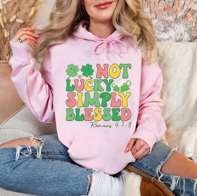 Puede incluir: Sudadera rosa con un gr&aacute;fico que dice "Not Lucky Simply Blessed" con tr&eacute;boles y jarras de cerveza. El texto "Romanos 4:7-8" est&aacute; debajo del gr&aacute;fico.