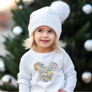 May include: A child wearing a white knitted hat with a pom-pom and a white sweatshirt. The sweatshirt has a colourful graphic of the Disneyland Resort logo in the shape of Mickey Mouse's head.