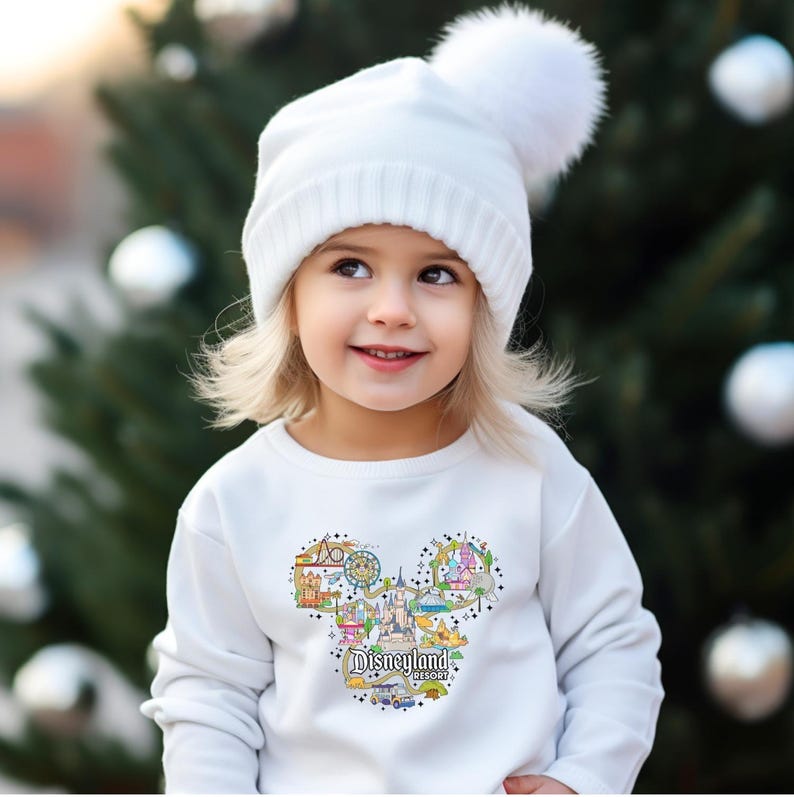 May include: A child wearing a white knitted hat with a pom-pom and a white sweatshirt. The sweatshirt has a colourful graphic of the Disneyland Resort logo in the shape of Mickey Mouse's head.