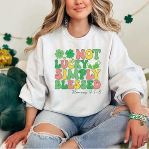 Puede incluir: Sudadera blanca con un gr&aacute;fico verde, amarillo y rosa que dice "Not Lucky Simply Blessed Romans 4:7-8".