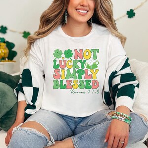 Puede incluir: Sudadera blanca con gr&aacute;ficos de tr&eacute;bol verde y el texto "NOT LUCKY SIMPLY BLESSED Romans 4:7-8".