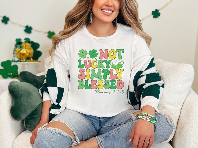 Puede incluir: Sudadera blanca con gr&aacute;ficos de tr&eacute;bol verde y el texto "NOT LUCKY SIMPLY BLESSED Romans 4:7-8".