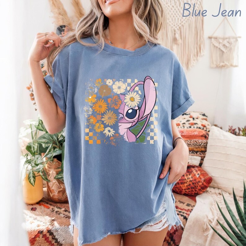 Puede incluir: Camiseta azul vaquero con un gr&aacute;fico de un personaje de dibujos animados y un dise&ntilde;o floral. El personaje es rosa y blanco con un ojo grande. El dise&ntilde;o floral incluye flores naranjas y blancas. El texto "Blue Jean" est&aacute; encima del gr&aacute;fico.