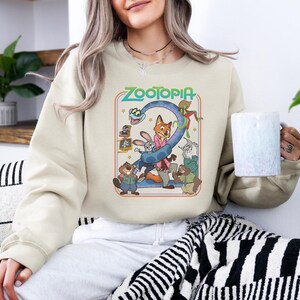 Pu&ograve; includere: Felpa color crema con una grafica colorata del film "Zootropolis". Il design include personaggi come Judy Hopps e Nick Wilde, con il titolo del film in un font retr&ograve;. La felpa &egrave; uno stile comodo e casual.