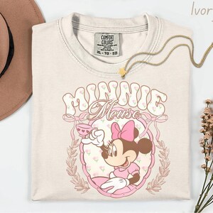 Puede incluir: Camiseta de color marfil con un gr&aacute;fico de Minnie Mouse. El dise&ntilde;o incluye el texto "Minnie Mouse" en una fuente floral, un lazo rosa y una ilustraci&oacute;n de dibujos animados de Minnie Mouse sosteniendo una taza de t&eacute;. Un collar de oro con un colgante de estrella est&aacute; sobre la camiseta.