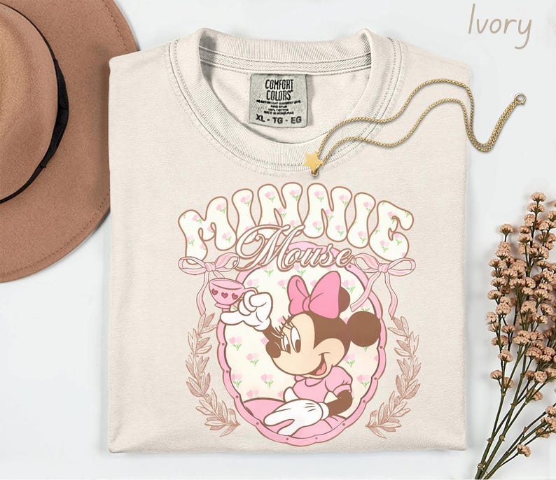 Puede incluir: Camiseta de color marfil con un gr&aacute;fico de Minnie Mouse. El dise&ntilde;o incluye el texto "Minnie Mouse" en una fuente floral, un lazo rosa y una ilustraci&oacute;n de dibujos animados de Minnie Mouse sosteniendo una taza de t&eacute;. Un collar de oro con un colgante de estrella est&aacute; sobre la camiseta.