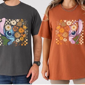 Puede incluir: Dos camisetas, una gris oscuro y otra color &oacute;xido, cada una con un dise&ntilde;o de personaje de dibujos animados. El dise&ntilde;o incluye un personaje con una corona de flores y elementos florales, sobre un fondo a cuadros.