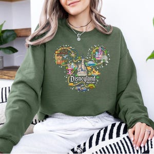 May include: An olive green sweatshirt with a Disneyland Resort graphic in the shape of Mickey Mouse's head. The graphic features illustrations of park attractions and the Disneyland Resort logo.