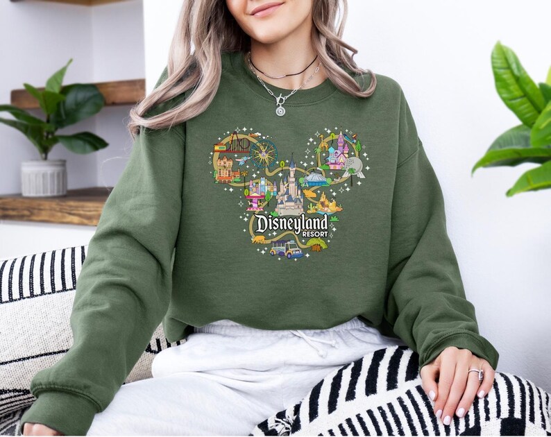 May include: An olive green sweatshirt with a Disneyland Resort graphic in the shape of Mickey Mouse's head. The graphic features illustrations of park attractions and the Disneyland Resort logo.