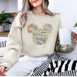 May include: A beige sweatshirt with a Disneyland Resort graphic in the shape of Mickey Mouse's head. The design includes the castle, rides, and the Disneyland Resort text. The model is holding a white mug. The sweatshirt is a neutral colour.