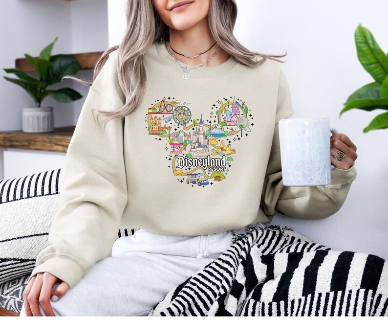 May include: A beige sweatshirt with a Disneyland Resort graphic in the shape of Mickey Mouse's head. The design includes the castle, rides, and the Disneyland Resort text. The model is holding a white mug. The sweatshirt is a neutral colour.