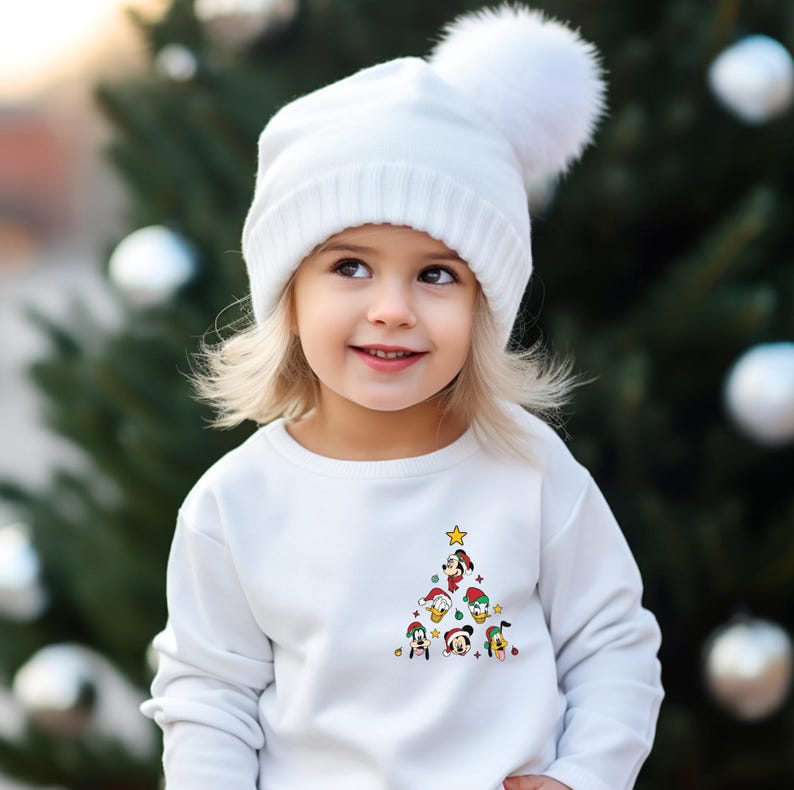 Pode incluir: Uma camisola branca com um desenho de &aacute;rvore de Natal com v&aacute;rios personagens da Disney, incluindo Mickey Mouse, Pato Donald, Pateta e Pluto. A &aacute;rvore est&aacute; decorada com enfeites e uma estrela no topo. A crian&ccedil;a est&aacute; usando um chap&eacute;u branco de malha com um pompom branco.