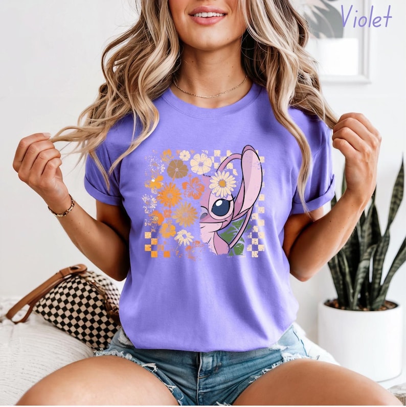 Puede incluir: Camiseta lila con un personaje de dibujos animados con una flor en la oreja, rodeado de un dise&ntilde;o floral en tonos naranja, amarillo y blanco. El personaje est&aacute; sobre un fondo a cuadros. La palabra "Violet" es visible.