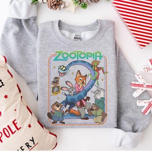 Pu&ograve; includere: Una felpa grigio chiaro con una grafica colorata del film "Zootropolis". La grafica include la parola "Zootropolis" in verde e blu, insieme a diversi personaggi animati. La felpa &egrave; circondata da decorazioni natalizie.
