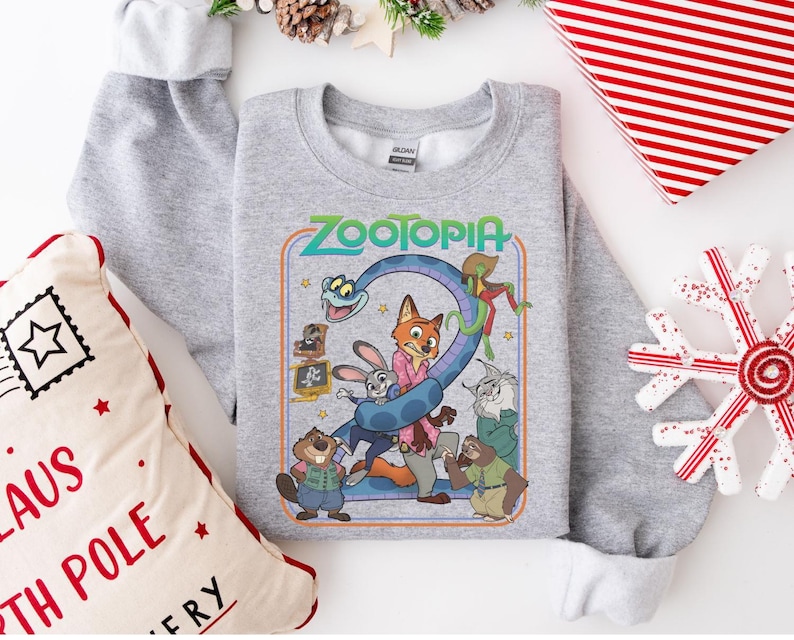 Pu&ograve; includere: Una felpa grigio chiaro con una grafica colorata del film "Zootropolis". La grafica include la parola "Zootropolis" in verde e blu, insieme a diversi personaggi animati. La felpa &egrave; circondata da decorazioni natalizie.