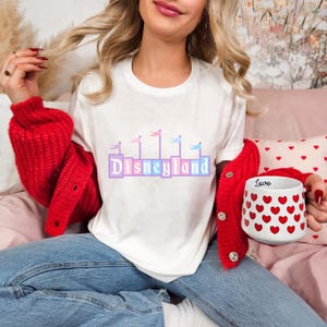 May include: A white t-shirt with a pink and blue Disneyland logo printed on the front. The person wearing the shirt is holding a white mug with red hearts.