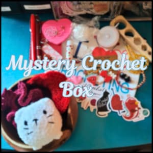 May include: A Mystery Crochet Box filled with yarn, crochet hooks, and other supplies. The box includes a crocheted cat, a red heart, and a variety of other items.