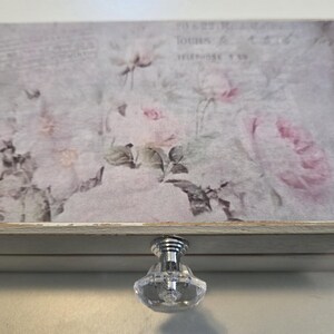 May include: A rectangular wooden box with a hinged lid featuring a faded floral print of pink roses and vintage script text. The box has a distressed white finish and a single clear, faceted crystal-style knob centered on the front edge.