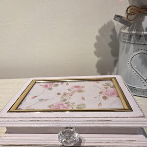 May include: A rectangular light pink wooden jewelry box featuring a glass top panel with a floral rose pattern. The box is framed with a thin gold-tone metal border and includes a single clear, faceted crystal-style knob on the front drawer.