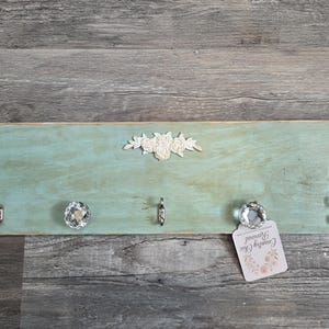 May include: A distressed, rectangular wooden wall-mounted key holder painted in a light teal color. It features four silver hooks, two crystal knobs, and a small decorative floral accent. A tag with text is attached.