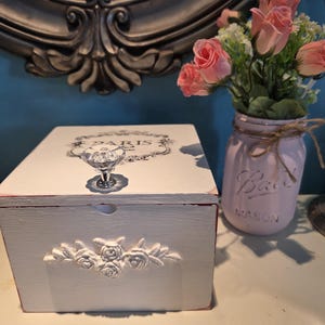 May include: A white wooden box with a crystal knob and a floral design. The box is decorated with the word "PARIS" and a decorative border. A pink mason jar with pink roses sits next to the box.