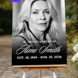 May include: A black and white memorial sign on a white easel. The sign features a portrait of a woman and the text "IN LOVING MEMORY OF Aime Smith OCT. 18, 1969 - AUG. 25, 2024". The Canva logo is in the upper left corner.