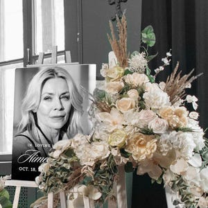 May include: A black and white memorial portrait of a woman with blonde hair, displayed on an easel. The portrait is surrounded by a floral arrangement of white, cream, and peach-colored flowers. The text "In Loving Memory Aime Oct. 18" is visible.