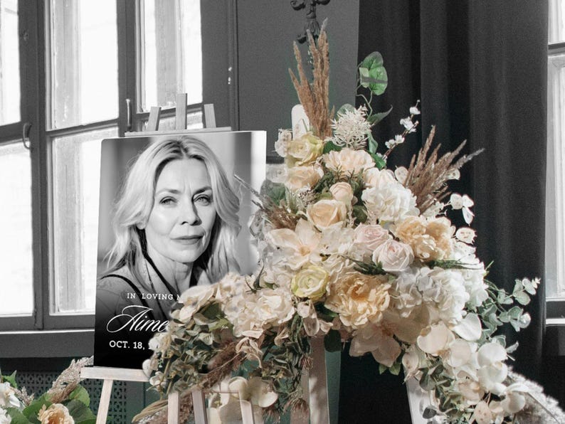 May include: A black and white memorial portrait of a woman with blonde hair, displayed on an easel. The portrait is surrounded by a floral arrangement of white, cream, and peach-colored flowers. The text "In Loving Memory Aime Oct. 18" is visible.