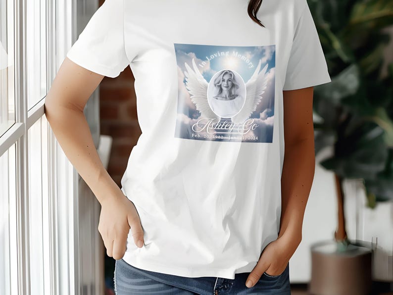 Puede incluir: Camiseta blanca con un dise&ntilde;o conmemorativo que presenta una foto de una mujer con alas de &aacute;ngel y el texto "In Loving Memory Ashley Go Feb. 20, 1988 - November 20, 2024"