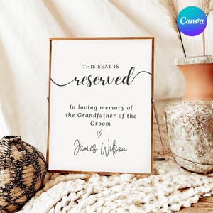 May include: A framed sign with the words "THIS SEAT IS reserved" in elegant script. Below, text reads "In loving memory of the Grandfather of the Groom" followed by a heart and a name. A vase and decorative sphere are in the background.